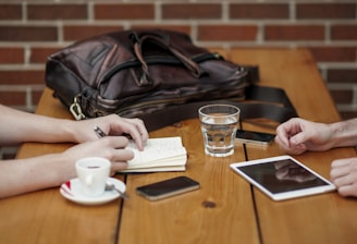 A wooden table hosts a casual meeting setup with two people present. One of them is taking notes in a small notebook with a pen in hand, while the other person has their hands resting on the table. A leather bag sits on the table next to a smartphone, a cup of coffee on a saucer, a glass of water, and a digital tablet.