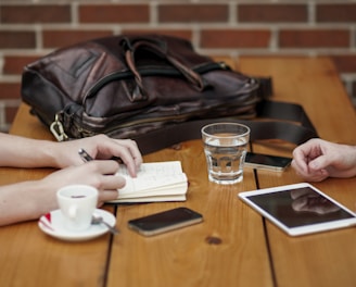 A wooden table hosts a casual meeting setup with two people present. One of them is taking notes in a small notebook with a pen in hand, while the other person has their hands resting on the table. A leather bag sits on the table next to a smartphone, a cup of coffee on a saucer, a glass of water, and a digital tablet.