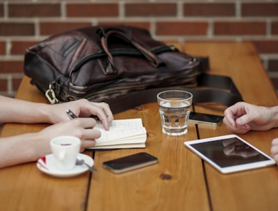 A wooden table hosts a casual meeting setup with two people present. One of them is taking notes in a small notebook with a pen in hand, while the other person has their hands resting on the table. A leather bag sits on the table next to a smartphone, a cup of coffee on a saucer, a glass of water, and a digital tablet.