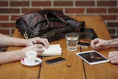 A wooden table hosts a casual meeting setup with two people present. One of them is taking notes in a small notebook with a pen in hand, while the other person has their hands resting on the table. A leather bag sits on the table next to a smartphone, a cup of coffee on a saucer, a glass of water, and a digital tablet.