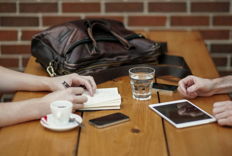 A wooden table hosts a casual meeting setup with two people present. One of them is taking notes in a small notebook with a pen in hand, while the other person has their hands resting on the table. A leather bag sits on the table next to a smartphone, a cup of coffee on a saucer, a glass of water, and a digital tablet.