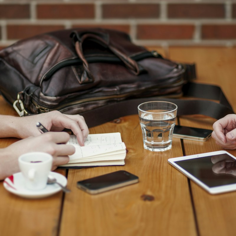 A wooden table hosts a casual meeting setup with two people present. One of them is taking notes in a small notebook with a pen in hand, while the other person has their hands resting on the table. A leather bag sits on the table next to a smartphone, a cup of coffee on a saucer, a glass of water, and a digital tablet.