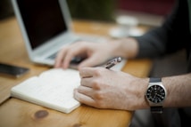 A person is writing in a notebook with a pen, sitting at a wooden table. A laptop and a smartphone are on the table, suggesting a work or study environment. The person is wearing a watch and the overall setting looks relaxed and focused.