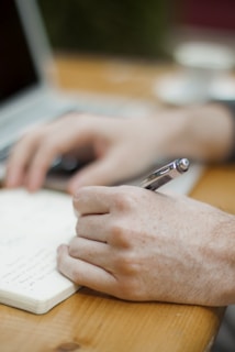 A person is writing in a notebook with a pen, sitting at a wooden table. A laptop and a smartphone are on the table, suggesting a work or study environment. The person is wearing a watch and the overall setting looks relaxed and focused.