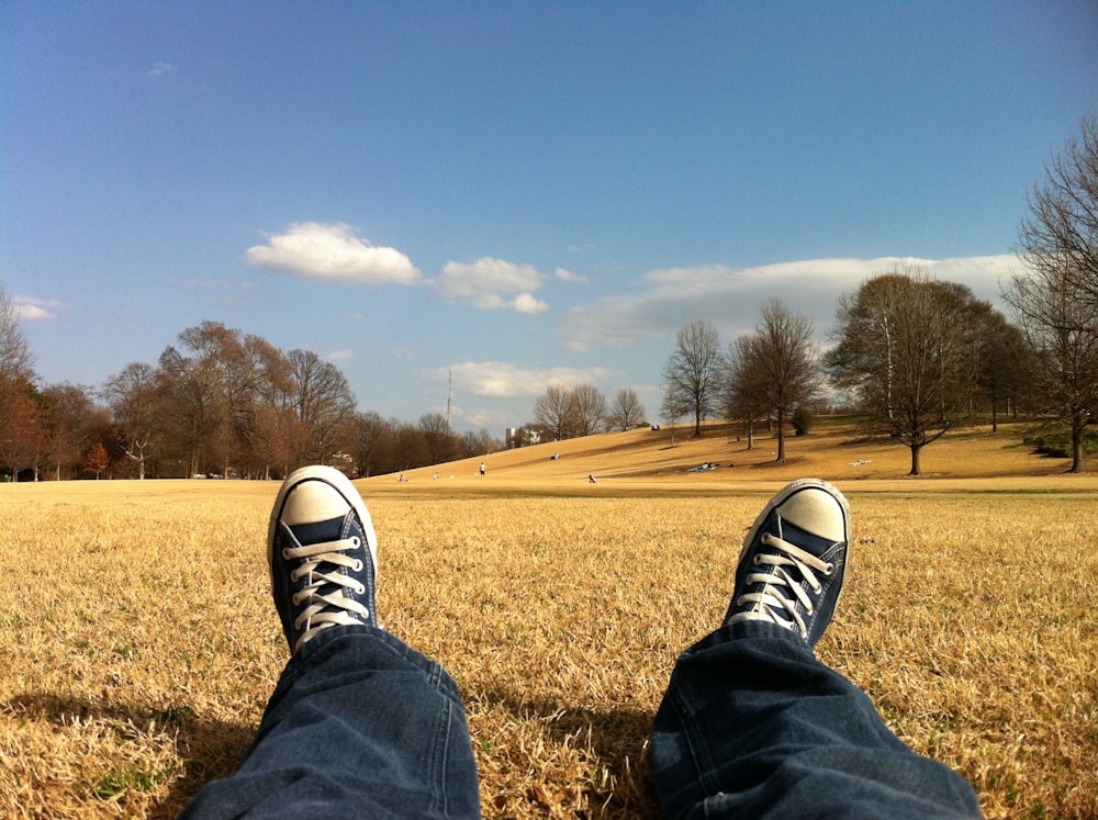 pessoa usando sapatos azuis e brancos Converse All Star enquanto está sentada na grama marrom durante o dia