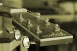 A close-up view of the headstock of an acoustic guitar with tuning pegs and strings visible. The image is in a sepia tone, giving it a vintage appearance. The guitar head features a brand logo.