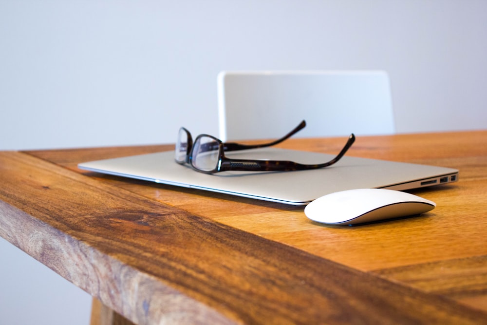 A pair of glasses on top of a laptop next to a mouse on a wooden surface