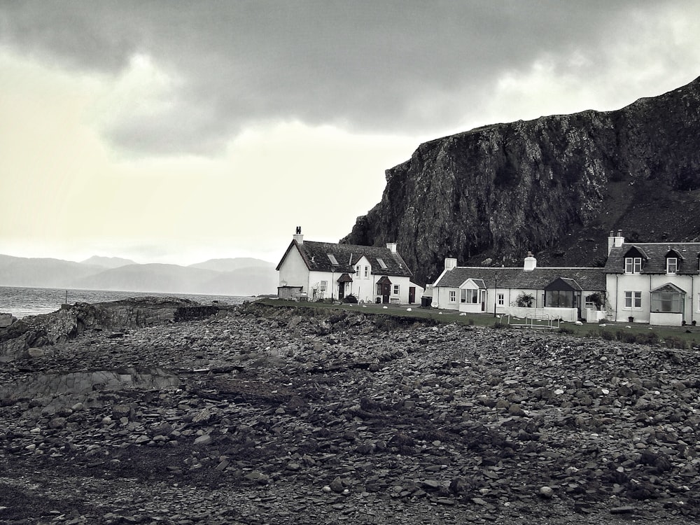 fotografia em tons de cinza de casas perto de montanhas rochosas e corpo d'água