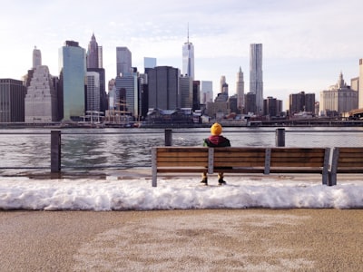 person in knit cap sitting on wooden bench in front of body of water during daytime faceless zoom background