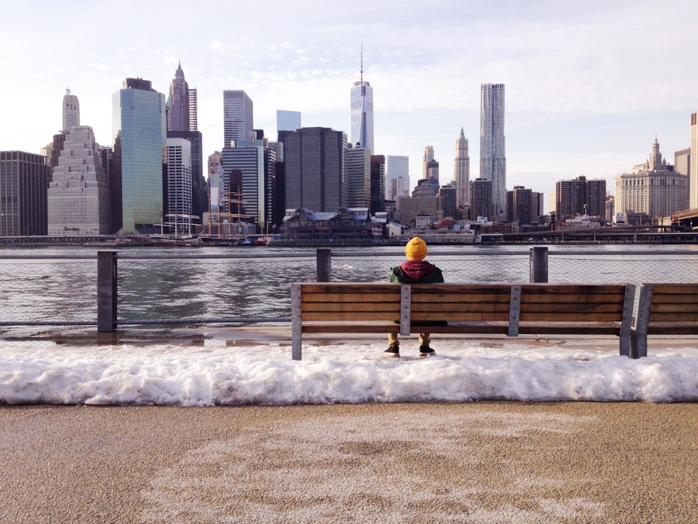 person in knit cap sitting on wooden bench in front of body of water during daytime