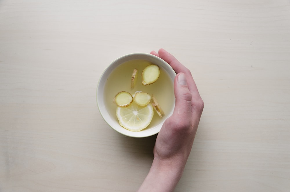 Personne tenant un bol blanc avec du citron vert tranché et du gingembre à l’intérieur