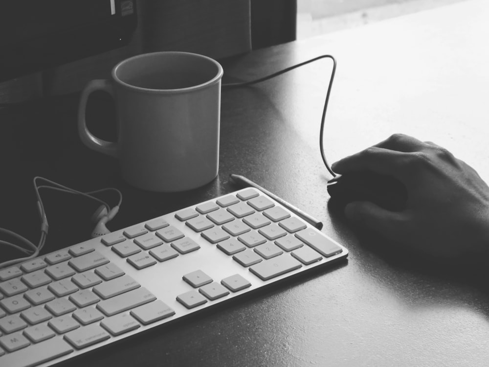 Graustufenfotografie einer Person, die eine Computermaus in der Nähe von Tastatur und Tasse hält