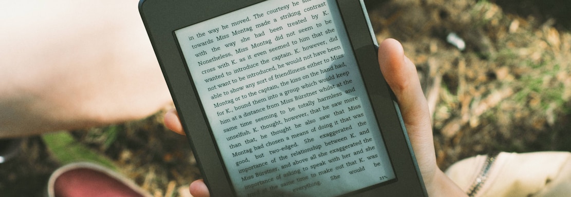 A person's hand holding a Kindle device displaying text. The Kindle is held outdoors, with part of their legs and the grass underneath visible in the background. The lighting suggests a sunny day, and there is a relaxed, leisurely atmosphere.
