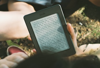 A person's hand holding a Kindle device displaying text. The Kindle is held outdoors, with part of their legs and the grass underneath visible in the background. The lighting suggests a sunny day, and there is a relaxed, leisurely atmosphere.