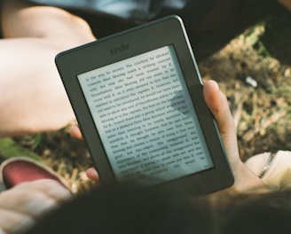 A person's hand holding a Kindle device displaying text. The Kindle is held outdoors, with part of their legs and the grass underneath visible in the background. The lighting suggests a sunny day, and there is a relaxed, leisurely atmosphere.