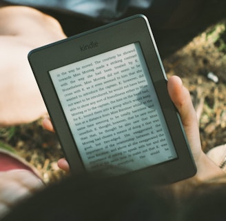 A person's hand holding a Kindle device displaying text. The Kindle is held outdoors, with part of their legs and the grass underneath visible in the background. The lighting suggests a sunny day, and there is a relaxed, leisurely atmosphere.