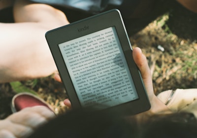 A person's hand holding a Kindle device displaying text. The Kindle is held outdoors, with part of their legs and the grass underneath visible in the background. The lighting suggests a sunny day, and there is a relaxed, leisurely atmosphere.