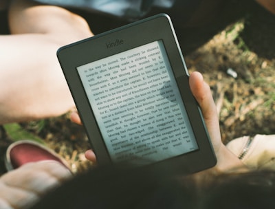 A person's hand holding a Kindle device displaying text. The Kindle is held outdoors, with part of their legs and the grass underneath visible in the background. The lighting suggests a sunny day, and there is a relaxed, leisurely atmosphere.
