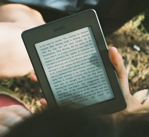 A person's hand holding a Kindle device displaying text. The Kindle is held outdoors, with part of their legs and the grass underneath visible in the background. The lighting suggests a sunny day, and there is a relaxed, leisurely atmosphere.