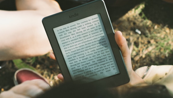A person's hand holding a Kindle device displaying text. The Kindle is held outdoors, with part of their legs and the grass underneath visible in the background. The lighting suggests a sunny day, and there is a relaxed, leisurely atmosphere.