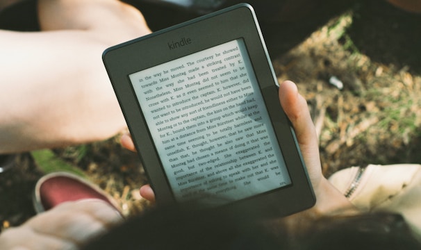 A person's hand holding a Kindle device displaying text. The Kindle is held outdoors, with part of their legs and the grass underneath visible in the background. The lighting suggests a sunny day, and there is a relaxed, leisurely atmosphere.