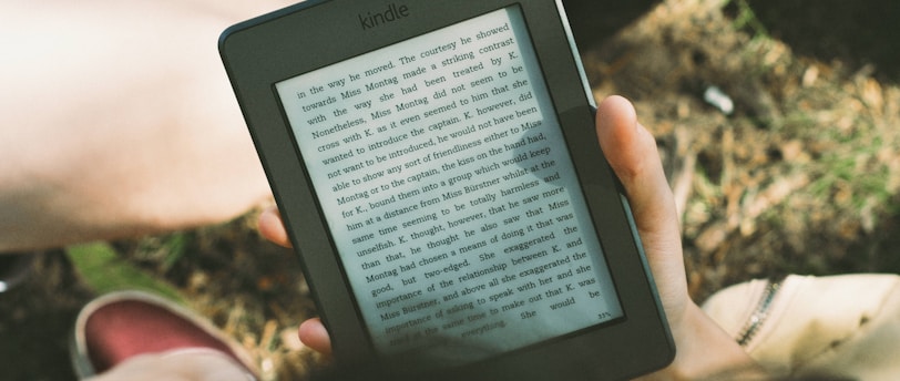 A person's hand holding a Kindle device displaying text. The Kindle is held outdoors, with part of their legs and the grass underneath visible in the background. The lighting suggests a sunny day, and there is a relaxed, leisurely atmosphere.