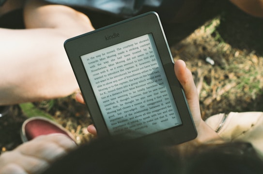 A person's hand holding a Kindle device displaying text. The Kindle is held outdoors, with part of their legs and the grass underneath visible in the background. The lighting suggests a sunny day, and there is a relaxed, leisurely atmosphere.