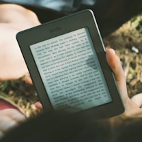 A person's hand holding a Kindle device displaying text. The Kindle is held outdoors, with part of their legs and the grass underneath visible in the background. The lighting suggests a sunny day, and there is a relaxed, leisurely atmosphere.