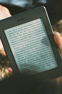 A person's hand holding a Kindle device displaying text. The Kindle is held outdoors, with part of their legs and the grass underneath visible in the background. The lighting suggests a sunny day, and there is a relaxed, leisurely atmosphere.