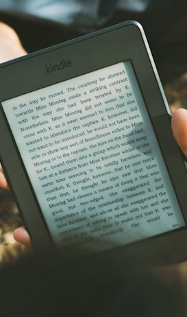A person's hand holding a Kindle device displaying text. The Kindle is held outdoors, with part of their legs and the grass underneath visible in the background. The lighting suggests a sunny day, and there is a relaxed, leisurely atmosphere.