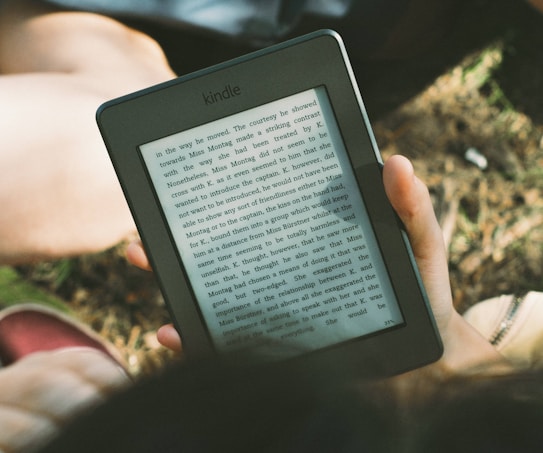 A person's hand holding a Kindle device displaying text. The Kindle is held outdoors, with part of their legs and the grass underneath visible in the background. The lighting suggests a sunny day, and there is a relaxed, leisurely atmosphere.