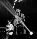 A band performs on stage with a singer playing an electric guitar at the center. A keyboard player stands to the left, concentrating on his instrument. Stage lights create a focused beam on the singer, adding dramatic effect. Smoke from a fog machine drifts to the right, enhancing the concert atmosphere.