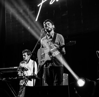 A band performs on stage with a singer playing an electric guitar at the center. A keyboard player stands to the left, concentrating on his instrument. Stage lights create a focused beam on the singer, adding dramatic effect. Smoke from a fog machine drifts to the right, enhancing the concert atmosphere.