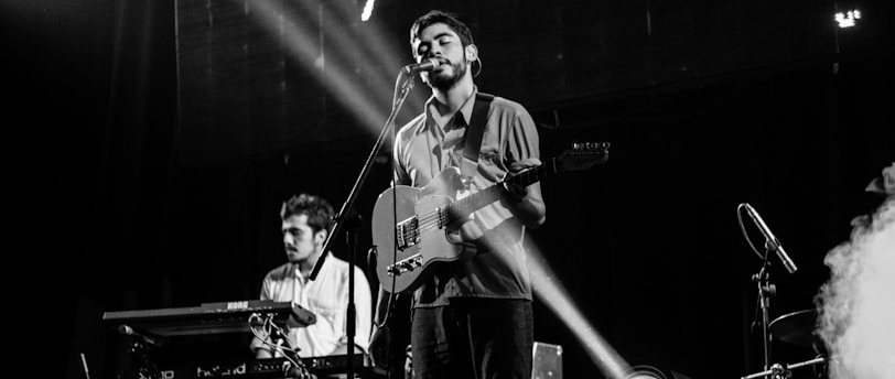 A band performs on stage with a singer playing an electric guitar at the center. A keyboard player stands to the left, concentrating on his instrument. Stage lights create a focused beam on the singer, adding dramatic effect. Smoke from a fog machine drifts to the right, enhancing the concert atmosphere.
