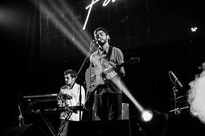 A band performs on stage with a singer playing an electric guitar at the center. A keyboard player stands to the left, concentrating on his instrument. Stage lights create a focused beam on the singer, adding dramatic effect. Smoke from a fog machine drifts to the right, enhancing the concert atmosphere.