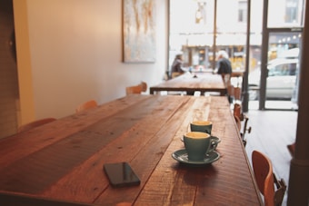 A wooden table in a cozy caf&eacute; with two colored coffee cups placed on it. There is a smartphone nearby, and in the background, two people are sitting and talking near a large window with a view of the street outside.