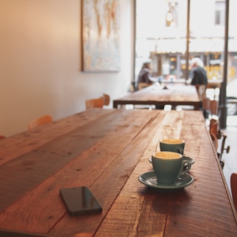 A wooden table in a cozy café with two colored coffee cups placed on it. There is a smartphone nearby, and in the background, two people are sitting and talking near a large window with a view of the street outside.