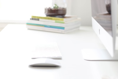 A sleek, modern desktop computer setup on a clean white desk. The image features a keyboard and mouse in focus, with a stack of books and a small glass item with a plant in the background.