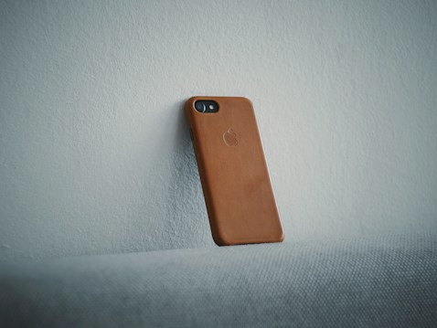 A smartphone with a brown leather case is leaning against a light-colored textured wall. The angle of the phone gives a minimalist and clean aesthetic. The logo on the case suggests it is designed for an Apple device.
