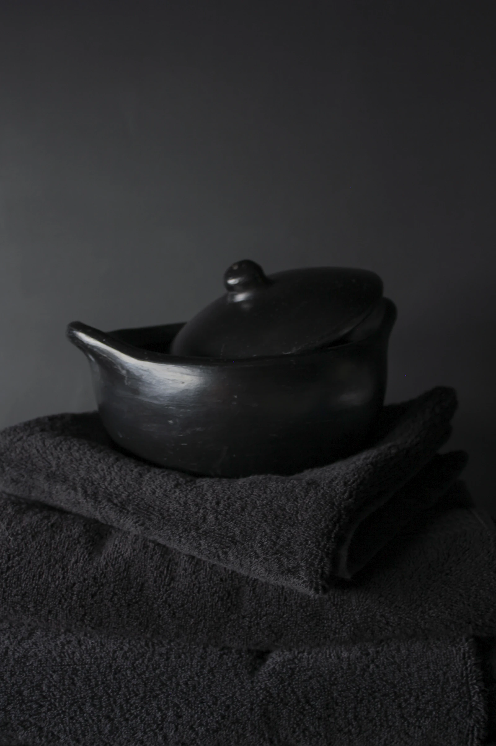 Matte black lidded pot resting on folded charcoal cloth against a dark wall