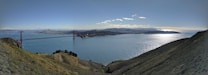 A panoramic view of a large body of water with the iconic Golden Gate Bridge stretching across it. The skyline of a city is visible in the distance, set against a backdrop of rolling hills and a partly cloudy sky. Sunlight glistens on the water's surface, creating a serene and expansive atmosphere.