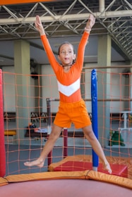 A young girl wearing an orange gymnastics outfit is enthusiastically jumping on a trampoline. Her arms are raised high, and she appears focused and joyful. The trampoline is surrounded by safety nets, and the setting looks like an indoor gymnasium with a high ceiling and several structural supports visible in the background.