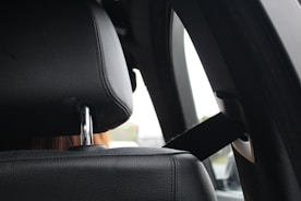 A close-up view of a black car seat headrest, showing the texture of the leather and the shiny metal support rods. Next to it is a seatbelt, which is attached to the car's interior. The background is slightly out of focus, hinting at a car's window with a light-colored exterior scene.