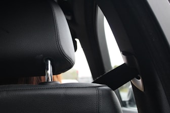 A close-up view of a black car seat headrest, showing the texture of the leather and the shiny metal support rods. Next to it is a seatbelt, which is attached to the car's interior. The background is slightly out of focus, hinting at a car's window with a light-colored exterior scene.