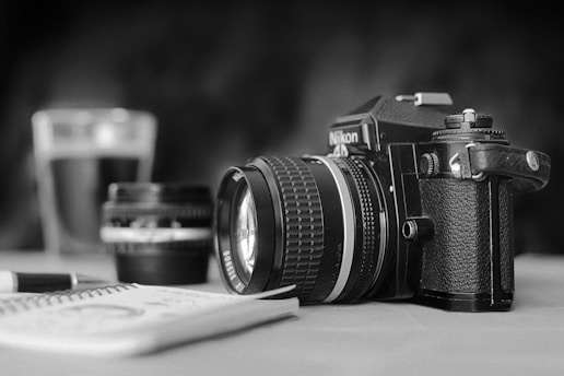A black and white photograph featuring a vintage Nikon camera with a prominent lens, placed on a table. In the background, a blurry camera lens and a glass can be seen, adding depth to the composition. A notebook with scribbles and a pen lie in the foreground, suggesting a moment of creativity or planning.