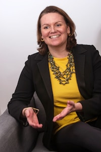 A woman is sitting on a gray chair, smiling and gesturing with her hands. She is wearing a black blazer over a bright yellow top and has a chunky necklace made of greenish-brown beads. Her short, brown hair is parted to the side, and she is accessorized with gold hoop earrings.
