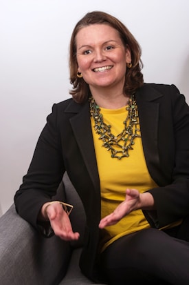 A woman is sitting on a gray chair, smiling and gesturing with her hands. She is wearing a black blazer over a bright yellow top and has a chunky necklace made of greenish-brown beads. Her short, brown hair is parted to the side, and she is accessorized with gold hoop earrings.