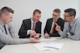 Four men in business attire are engaged in a discussion around a table. One man appears to be explaining something while pointing at some documents in front of them. The others are listening attentively, and one holds a pen and a mobile device. The setting is a plain office environment.