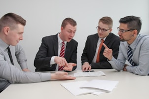 Four men in business attire are engaged in a discussion around a table. One man appears to be explaining something while pointing at some documents in front of them. The others are listening attentively, and one holds a pen and a mobile device. The setting is a plain office environment.