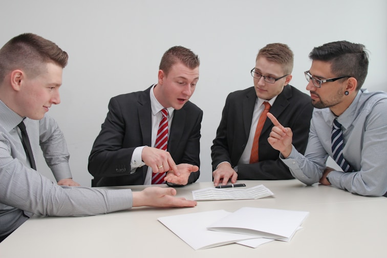 Four men in business attire are engaged in a discussion around a table. One man appears to be explaining something while pointing at some documents in front of them. The others are listening attentively, and one holds a pen and a mobile device. The setting is a plain office environment.